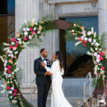 A couple dressed in formal attire pose beneath a flower arch on the terrace of Savanna Hall.