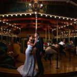 A couple in formal attire pose looking at each other on the carousel at night.