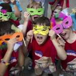 A group of excited kids, wearing Safari Day Camp shirts featuring a gorilla, show off the animal themed opera masks they