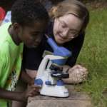 A kid looks through a microscope while an instructor looks one at Safari Camp.