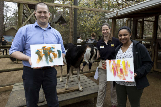 A group of two people and an Animal Care professional pose for a photo with a goat and animal created painting.
